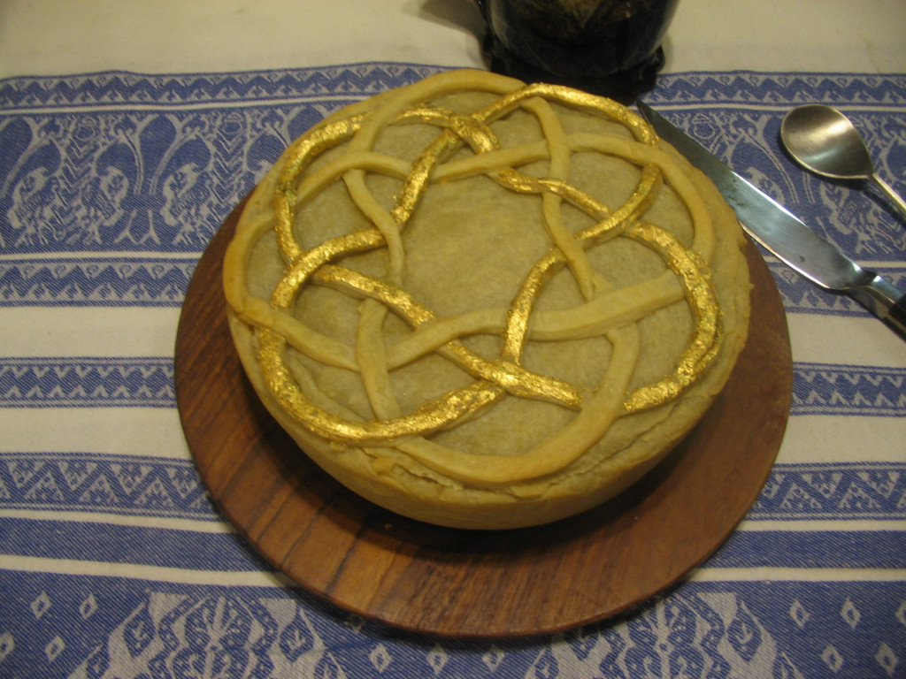 Overhead photo of pie on a wooden plate. Pie has a Celtic knotwork design in pastry on top, partially decorated with gold leaf.